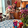 woman, cat, party_hat, kitchen, table, flowers, piñata, birthday_cake, candle, window, cabinet, glasses, tattoo, smile, dress, vase, food, indoor, celebration, happy