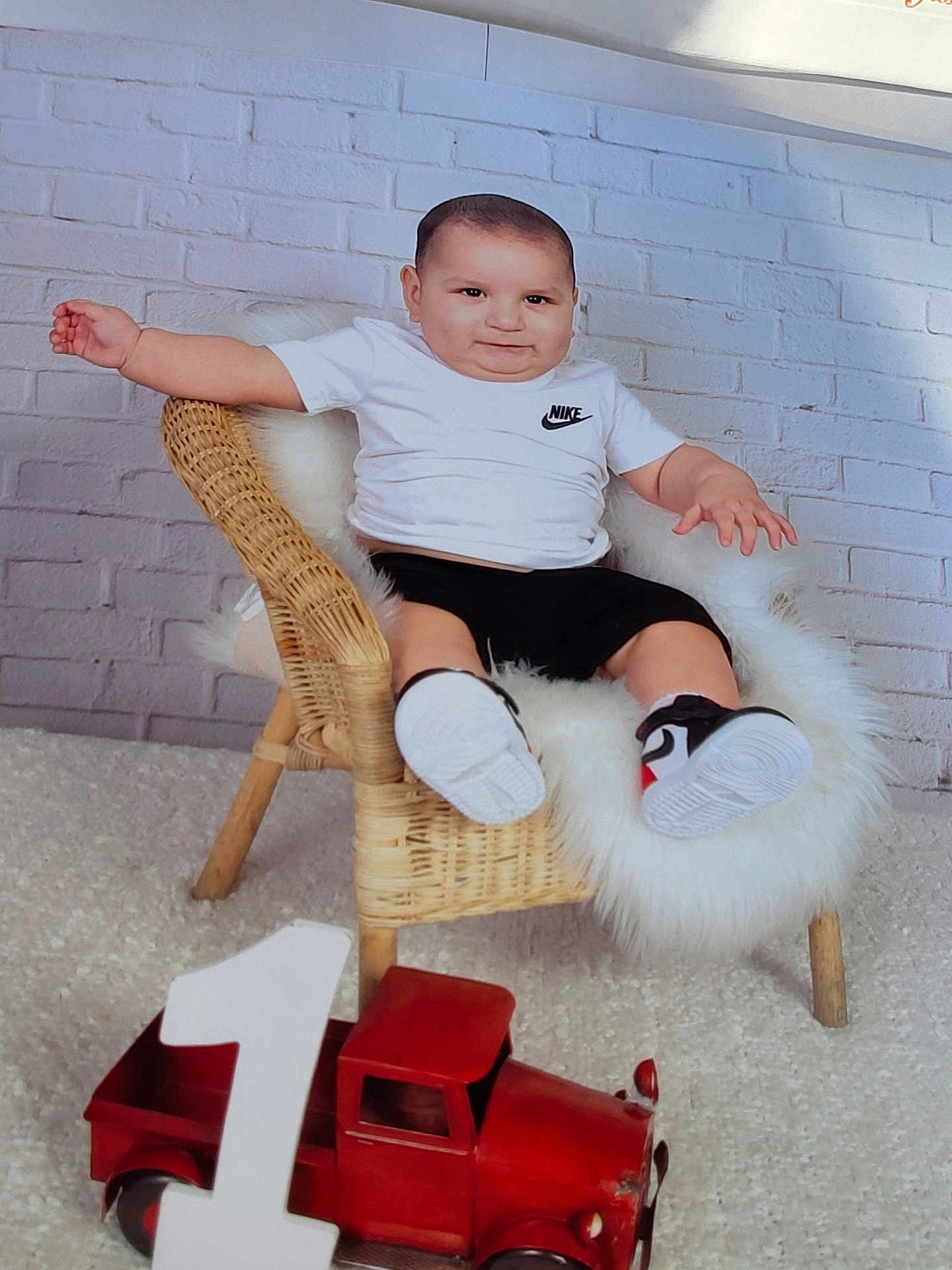 Elezar is registered to the contest to win money with this photo: black_shorts, brick_wall, carpet, celebration, child, cute, first_birthday, floor, happy, indoor, nike_logo, number_one, portrait, smiling, sneakers, toddler, toy_truck, white_fur, white_shirt, wicker_chair