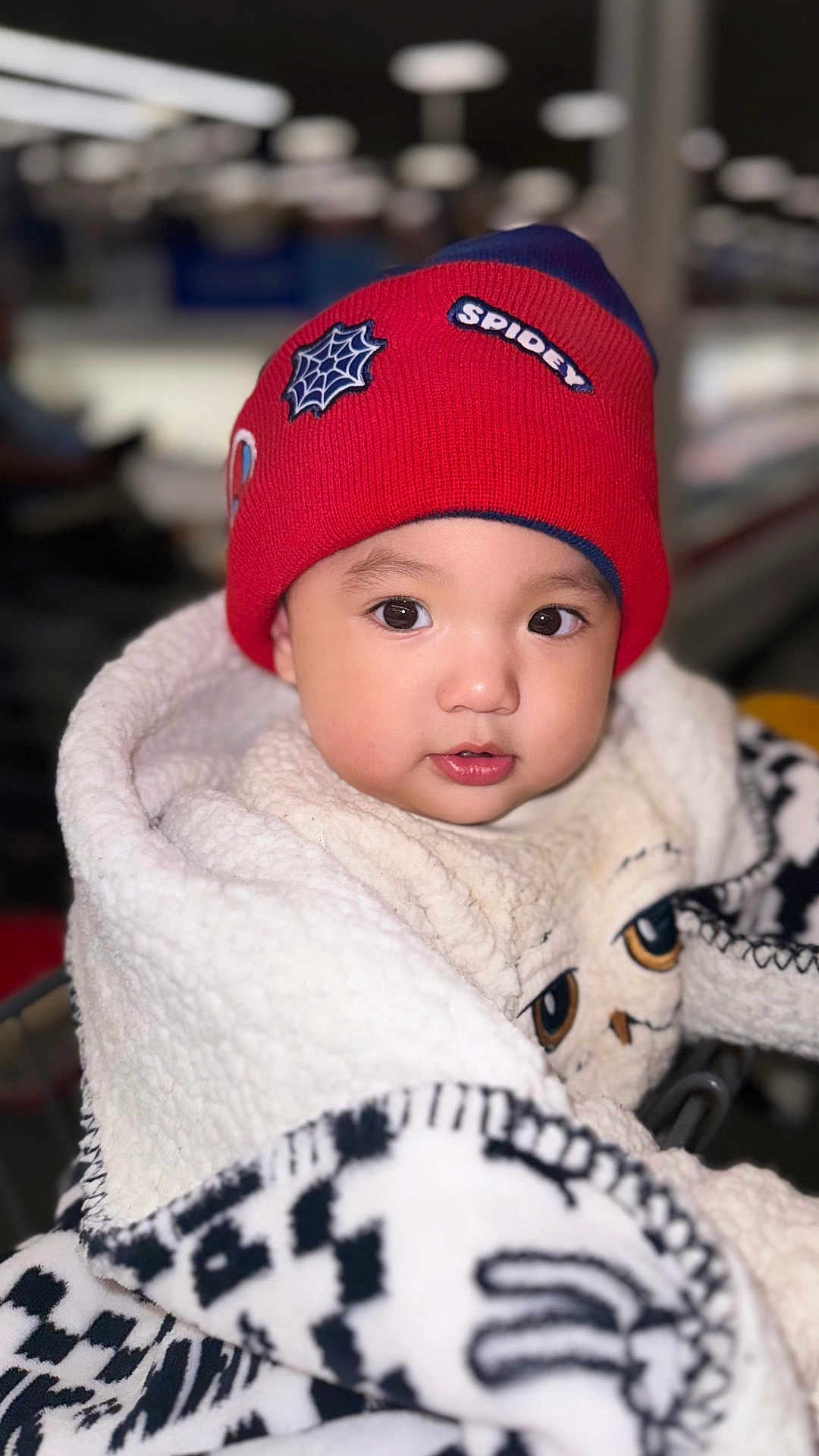 Drake is registered to the contest to win money with this photo: baby, child, red_beanie, hat, spiderman, blanket, fleece, indoors, portrait, cute, face, eyes, warm_clothing, winter_wear, soft_texture, young_child, cozy, closeup, apparel, infant