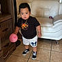 toddler, child, balloon, black_tshirt, dinosaur_print, striped_shorts, cartoon_emblem, black_sneakers, living_room, white_couch, wooden_furniture, tile_floor, standing, indoor, person, cute, young_child, casual_clothing, playful, bracelet