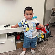 Drake is registered to the contest to win money with this photo: toddler, child, indoor, carpet, toy, socks, shorts, tshirt, remote_control, furniture, shoes, backpack, white_wall, cable, expression, person, standing, room, play, curious