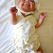 Mila is registered to the contest to win money with this photo: baby, infant, smiling, hat, white, lemon_pattern, clothing, bed, blanket, happy, cute, child, fist, lying_down, cheeks, playful, skin, person, infant_clothing, indoors
