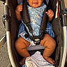 baby, stroller, child, smiling, blue_dress, hair_bun, blanket, sitting, outdoor, pavement, safety_harness, infant, cute, happy, person, footwear, black_hair, toddler, baby_clothing, seat