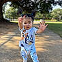 toddler, child, pigtails, outdoor, sunlight, cartoon_clothing, playful_pose, sidewalk, tree, greenery, shoes, casual_wear, young_child, person, cute, smiling, daylight, nature, fun, park