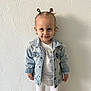 Camélia participe au concours pour gagner de l'argent avec cette photo : toddler, child, denim_jacket, white_clothing, standing, smile, hair_tied, pony_tail, blue_eyes, earrings, wall, textured_wall, portrait, indoor, cute, happy, person, fashion, casual_wear, young_child