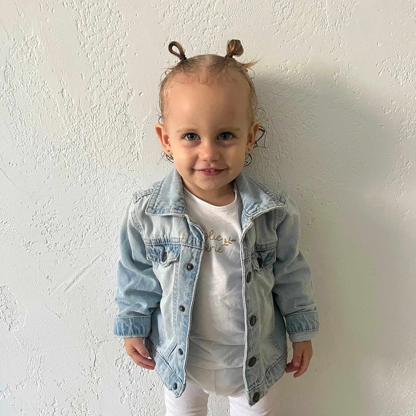 Camélia participe au concours pour gagner de l'argent avec cette photo : blue_eyes, casual_wear, child, cute, denim_jacket, earrings, fashion, hair_tied, happy, indoor, person, pony_tail, portrait, smile, standing, textured_wall, toddler, wall, white_clothing, young_child