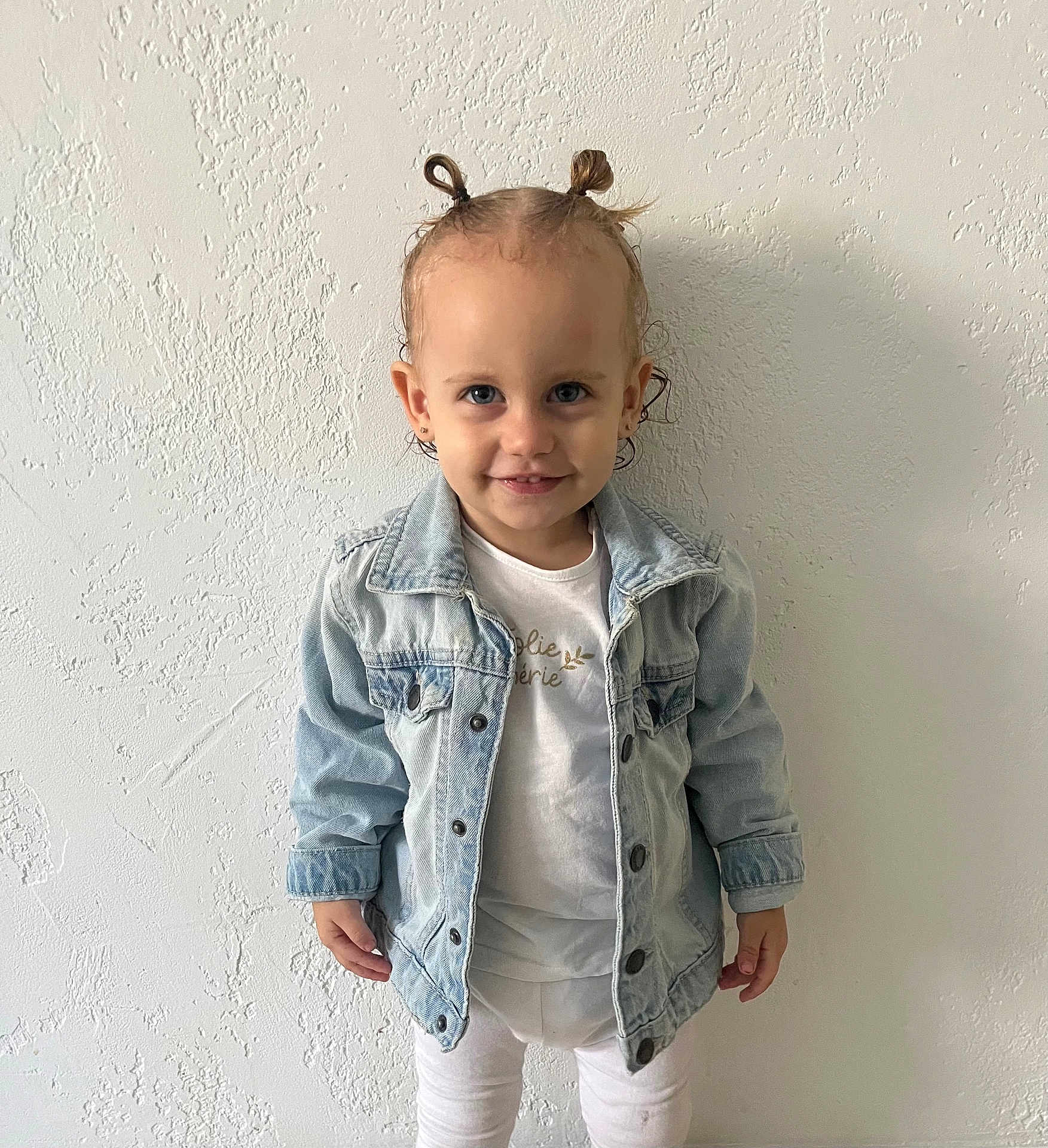 Camélia participe au concours pour gagner de l'argent avec cette photo : toddler, child, denim_jacket, white_clothing, standing, smile, hair_tied, pony_tail, blue_eyes, earrings, wall, textured_wall, portrait, indoor, cute, happy, person, fashion, casual_wear, young_child