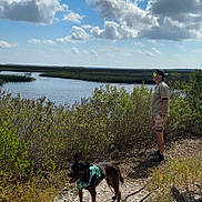 Paisley is registered to the contest to win money with this photo: dog, person, outdoor, nature, water, sky, clouds, trail, greenery, harness, sunlight, landscape, walking, leash, bushes, summer, casual_clothing, hat, shoes, exploring