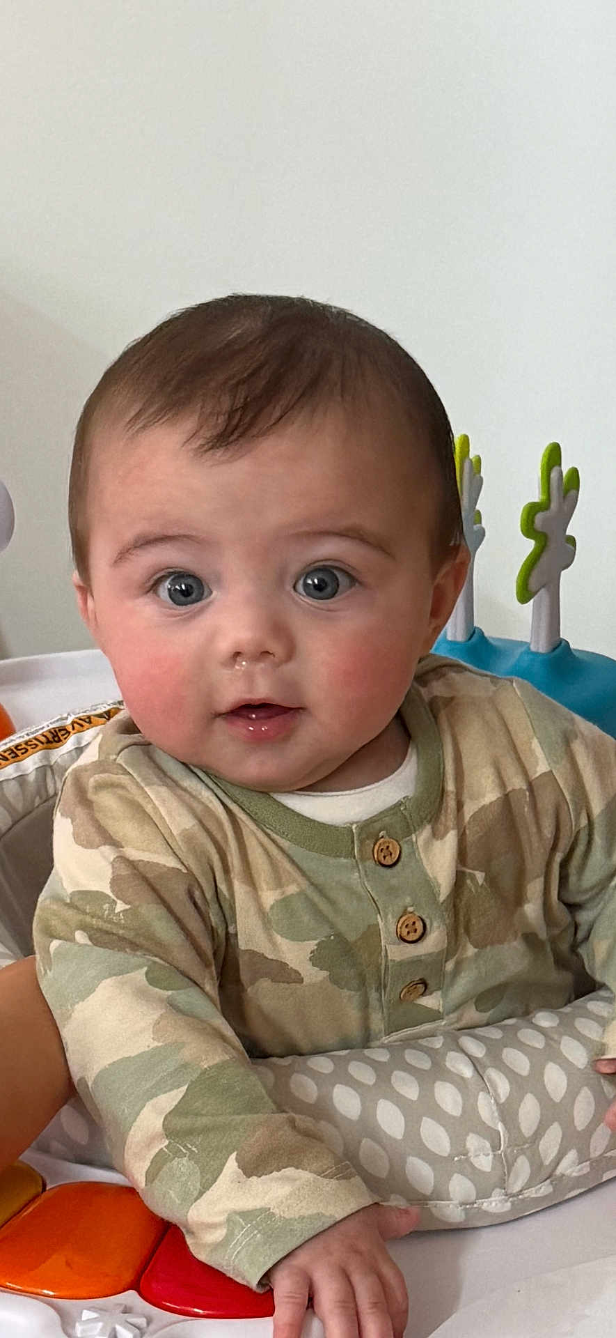 Ezekiel is registered to the contest to win money with this photo: baby, child, infant, face, eyes, rosy_cheeks, camo_clothing, onesie, hand, toy, activity_center, colorful, curious, indoor, portrait, sitting, playful, young_child, cute, expression