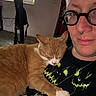 Franklin is registered to the contest to win money with this photo: cat, person, glasses, indoors, sleeping_cat, orange_cat, black_shirt, neon_green, pumpkin_face, nose_ring, closeup, affection, resting, cute, pet, casual, portrait, home, comfort, relaxation