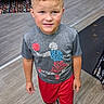 child, boy, curly_hair, tshirt, shorts, sneakers, floor, indoor, living_room, carpet, chair, cushion, table, smiling, standing, person, casual, young, curious, home