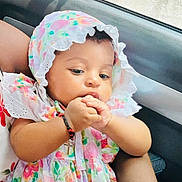 Kerri is registered to the contest to win money with this photo: baby, child, car, window, floral_dress, bonnet, hand, adult_arm, sitting, cute, infant, skin, clothing, portrait, indoors, seat, looking, person, young, support