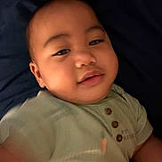 Tamasa is registered to the contest to win money with this photo: baby, infant, child, portrait, close_up, face, sleepy, calm, looking_at_camera, dark_background, blanket, bed, indoor, cozy, green_shirt, buttons, sitting, soft_focus, warm_lighting, cute