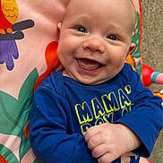 Asher is registered to the contest to win money with this photo: baby, smile, happy, infant, blue_shirt, text_on_clothing, colorful_mat, cartoon_bird, hand, face, eyes, cheeks, mouth, tongue, indoor, carpet, clothing, cute, child, portrait