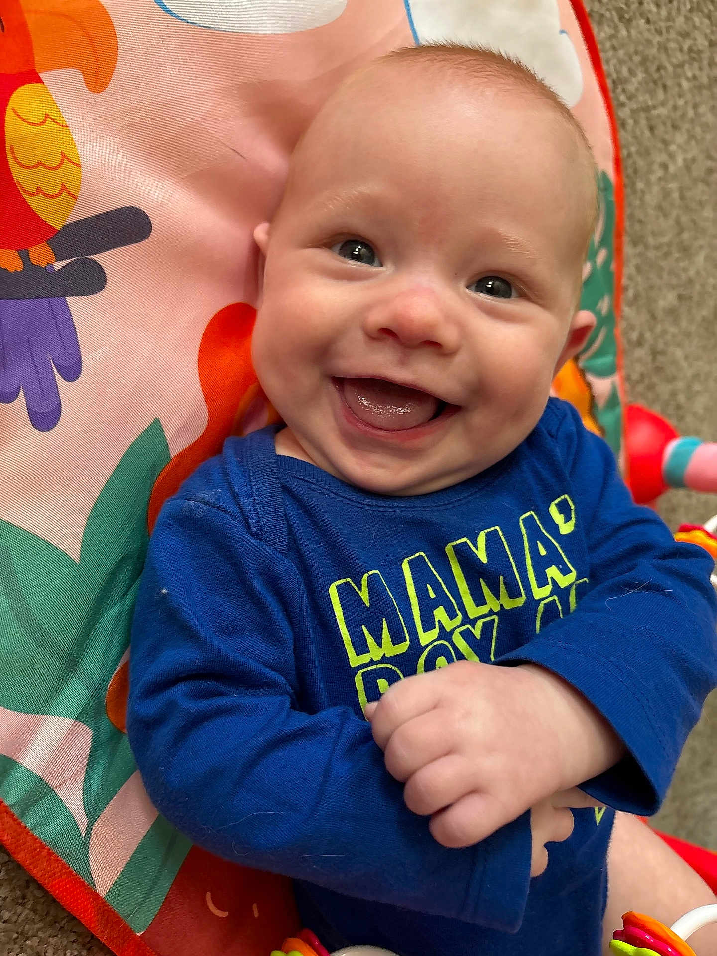 Asher is registered to the contest to win money with this photo: baby, smile, happy, infant, blue_shirt, text_on_clothing, colorful_mat, cartoon_bird, hand, face, eyes, cheeks, mouth, tongue, indoor, carpet, clothing, cute, child, portrait