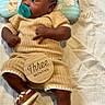baby, infant, pacifier, knitted_clothing, brown_shoes, wooden_sign, three_months, blanket, pillow, cartoon_character, lying_down, cute, child, person, indoors, soft_lighting, portrait, smiling, cozy, newborn