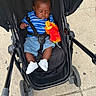 baby, child, stroller, toy, plush_toy, blue_shirt, striped, shorts, white_shoes, concrete, sidewalk, outdoor, person, infant, seatbelt, casual_clothing, black_stroller, walking, summer, cute