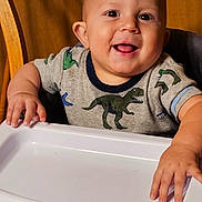 Zayriee is registered to the contest to win money with this photo: baby, child, clothing, cute, dinosaur_shirt, face, hands, happy, high_chair, indoor, infantino, person, playful, portrait, sitting, smiling, table, toddler, wooden_background, young