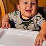 baby, child, clothing, cute, dinosaur_shirt, face, hands, happy, high_chair, indoor, infantino, person, playful, portrait, sitting, smiling, table, toddler, wooden_background, young