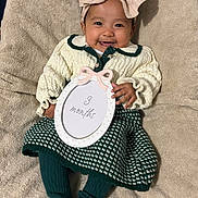 Emilia is registered to the contest to win money with this photo: baby, infant, smiling, bow, headband, knitted_sweater, dress, tights, socks, blanket, sign, portrait, cute, happy, child, person, indoor, soft_texture, cozy, three_months