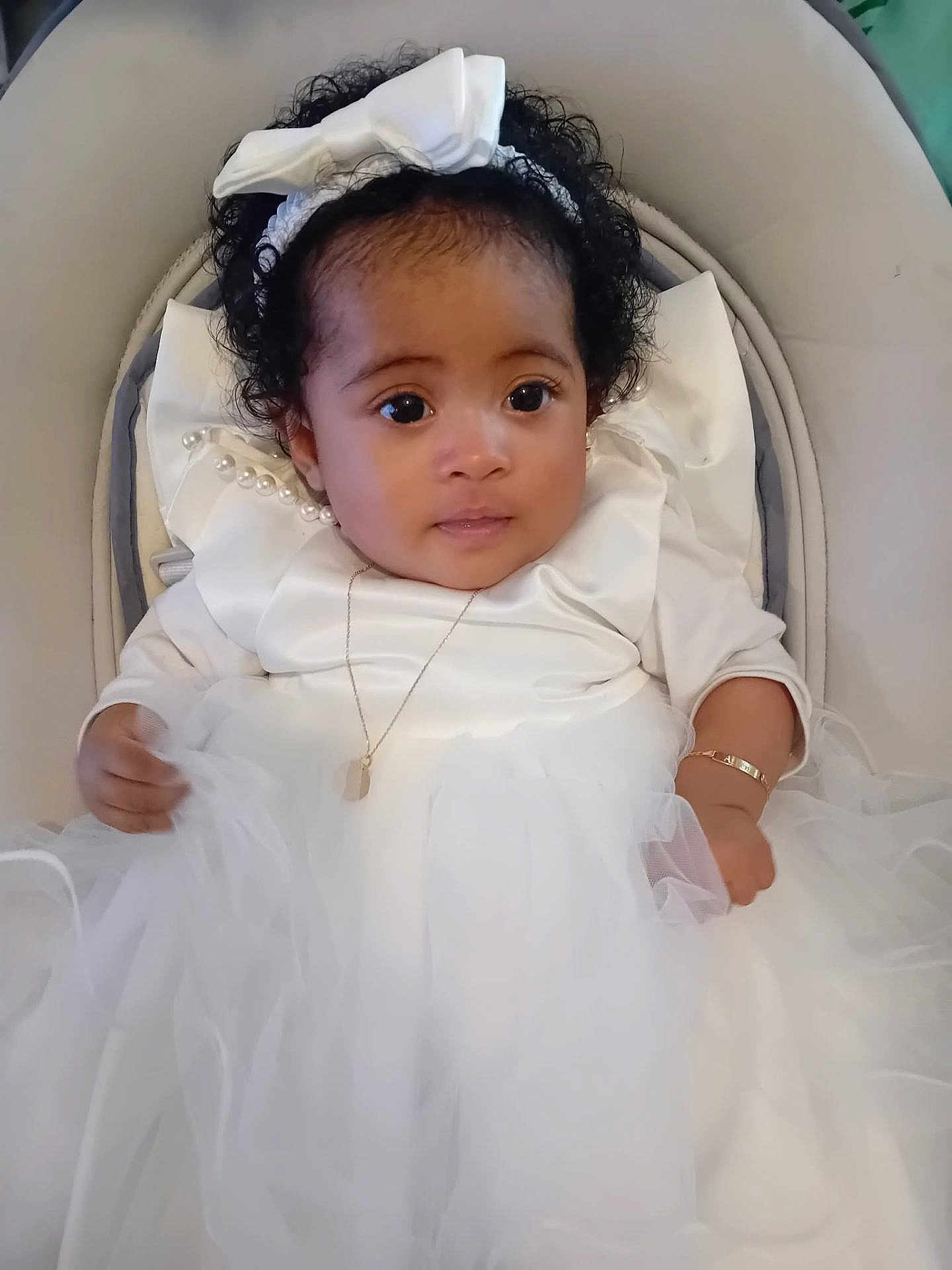 Athéna participe au concours pour gagner de l'argent avec cette photo : baby, infant, toddler, white_dress, bow_headband, curly_hair, big_eyes, necklace, bracelet, tulle_skirt, seated, stroller_seat, portrait, adorable, formal_outfit, pearl_detail, soft_lighting, closeup, smiling, baby_chair