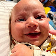Kaiden is registered to the contest to win money with this photo: baby, smiling, close_up, face, infant, child, joy, happy, patterned_clothing, pillow, striped, hand, skin, indoors, background, colorful, cute, portrait, young, expression