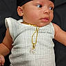 baby, infant, clothing, onesie, calvin_klein, cap, gold_chain, pendant, face, skin, laying_down, looking_sideways, cute, newborn, child, fashion, accessory, portrait, indoors, black_background