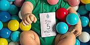 Lyam a rejoint le concours — aidez-le/la à gagner de superbes lots ! baby, child, infant, ball_pit, colorful_balls, green_onesie, milestone_card, smiling, sitting, playtime, cute, indoors, happy, skin, feet, hands, face, head, toes, portrait