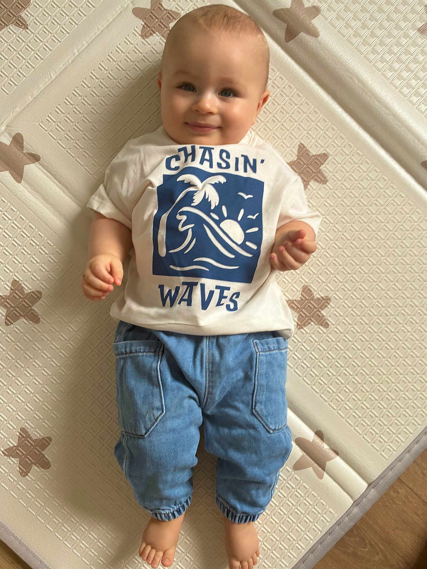 Lyam participe au concours pour gagner de l'argent avec cette photo : baby, child, smiling, tshirt, blue_pants, barefoot, floor_mat, star_pattern, indoor, person, cute, infant, happy, portrait, clothing, baby_clothes, head, feet, light_skin, casual