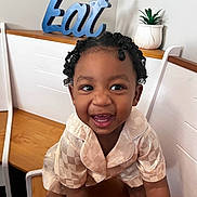 Jojo is registered to the contest to win money with this photo: toddler, child, smile, curly_hair, indoor, bench, wood, potted_plant, decor, sign, happy, clothing, person, face, cute, playful, table, furniture, expression, young