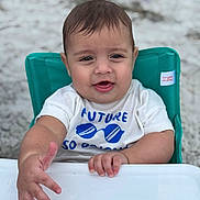 Alan joined the competition — help win amazing prizes! baby, child, high_chair, white_shirt, blue_text, sunglasses_graphic, hand, face, wet_hair, teal_chair, beach, sand, outdoor, person, infant, cute, smiling, portrait, summer, happy
