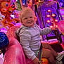 toddler, child, amusement_ride, pink_car, smiling, curly_hair, toy, steering_wheel, indoor, colorful, lights, carnival, fun, playful, cartoon, seatbelt, casual_clothing, hoodie, shoes, happy