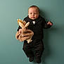 baby, infant, child, portrait, suit, bow_tie, stuffed_animal, plush_toy, teal_background, studio_lighting, lying_down, cute, adorable, formalwear, one_person, small_hands, soft_texture, neutral_expression, fashionable, toy_holder