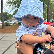 Jahlani is registered to the contest to win money with this photo: baby, child, hat, blue_hat, striped_shirt, tattoo, arm, outdoor, trees, parking_lot, person, holding, cute, curious, infant, casual_clothing, daytime, nature, portrait, expression