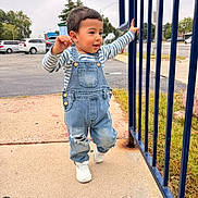 Nathan is registered to the contest to win money with this photo: child, toddler, boy, outdoor, portrait, walking, standing, smiling, striped_shirt, denim_overalls, sneakers, blue_fence, sidewalk, street, cars, trees, neighborhood, daylight, fashion, playful