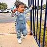 child, toddler, boy, outdoor, portrait, walking, standing, smiling, striped_shirt, denim_overalls, sneakers, blue_fence, sidewalk, street, cars, trees, neighborhood, daylight, fashion, playful