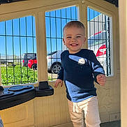 Tiago participe au concours pour gagner de l'argent avec cette photo : toddler, child, smiling, playhouse, blue_sweater, light_pants, sandals, sunlight, window, car, greenery, outdoor, daylight, happy, playing, young_child, portrait, person, cute, casual_clothing