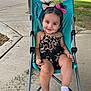toddler, child, smiling, stroller, outdoor, sidewalk, leopard_print, hair_bows, white_socks, happy, cute, seatbelt, greenery, concrete, playful, young_child, portrait, baby, casual, sunlight