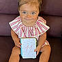baby, child, girl, blue_eyes, pink_bows, striped_outfit, sitting, couch, card, holding, smiling, shoes, legs, face, hair, indoors, cute, portrait, young_child, happy