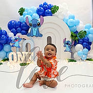Josias is registered to the contest to win money with this photo: baby, balloons, birthday, blue, celebration, child, clapping, cute, decorations, event, happy, indoor, lights, orange_shirt, party, photography, sitting, smiling, stitch, studio