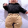 baby, infant, child, black_shirt, beige_pants, white_quilt, footwear, lying_down, cute, face, skin, hand, curious, expression, indoor, clothing, portrait, one_person, young, casual