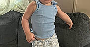 Hadley is registered to the contest to win money with this photo: toddler, child, baby, smiling, standing, portrait, indoor, couch, cushion, curly_hair, blue_top, shorts, clothing, curtain, window, home, furniture, happy, playful, photography