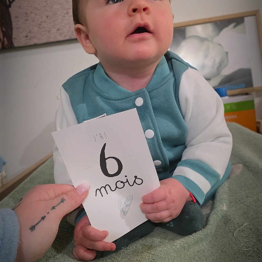 Loussio participe au concours pour gagner de l'argent avec cette photo : 6_months, baby, blanket, card, child, curious, cute, face, hand, holding, indoors, infant, person, photography, portrait, sitting, tattoo, teal_jacket, white_sleeves, young_child