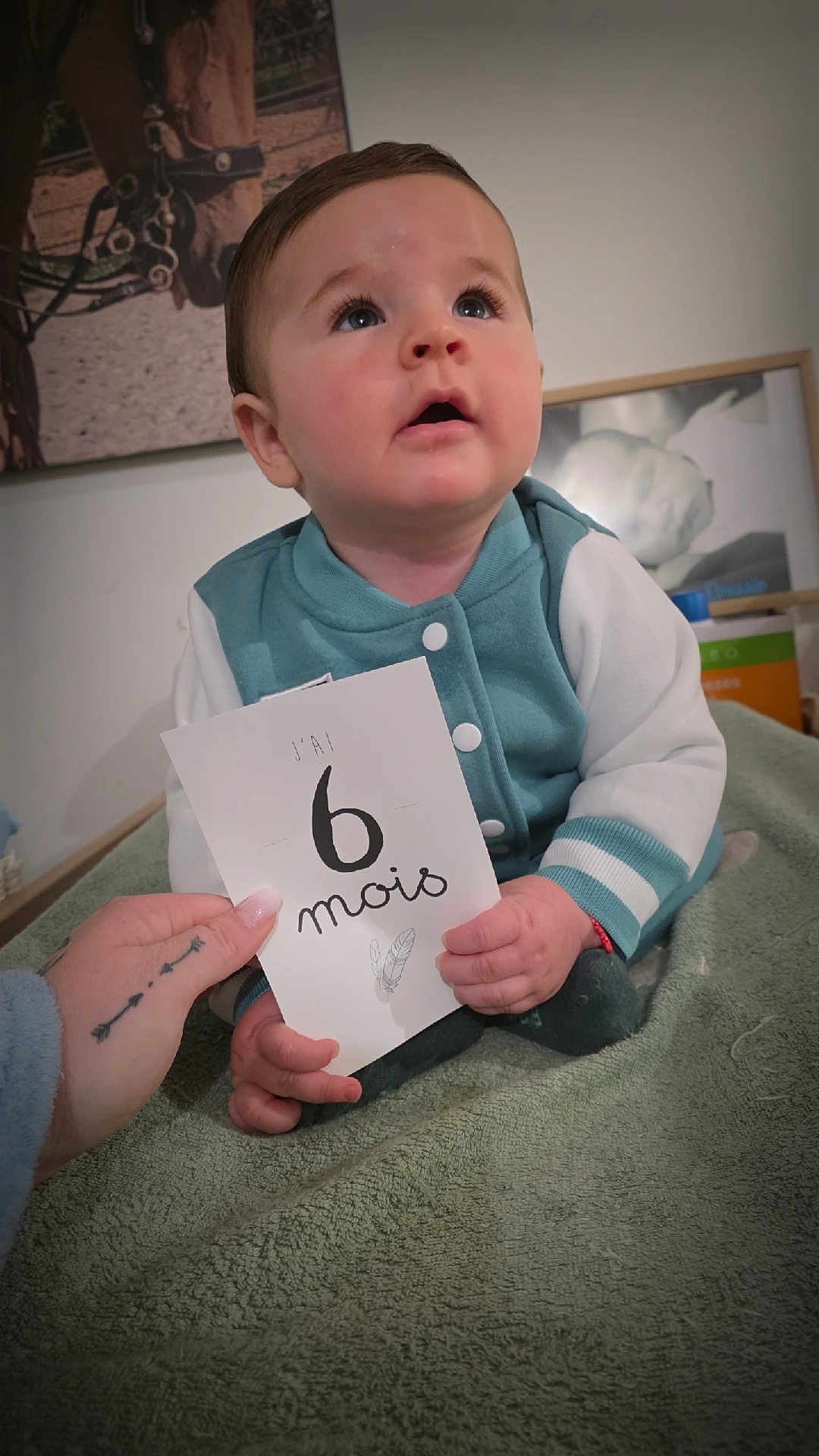 Loussio participe au concours pour gagner de l'argent avec cette photo : 6_months, baby, blanket, card, child, curious, cute, face, hand, holding, indoors, infant, person, photography, portrait, sitting, tattoo, teal_jacket, white_sleeves, young_child