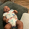Kieaie is registered to the contest to win money with this photo: baby, infant, onesie, dinosaur_pattern, couch, furniture, wooden_floor, baby_chair, indoor, person, child, cute, relaxed, skin, smiling, happy, legs, hands, head, casual