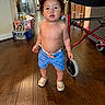 bare_chest, blue_shorts, cap, casual, child, curly_hair, expression, flooring, footwear, furniture, home, indoor, light, person, standing, toddler, toy, walker, wooden_floor, young_child