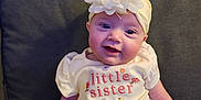 Ava is registered to the contest to win money with this photo: apparel, baby, child, couch, cute, flower_headband, furniture, hand, happy, indoor, infant, little_sister_text, person, portrait, seated, skin, smiling, tattoo, white_shirt, yellow_pants