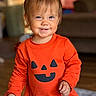 toddler, child, baby, smiling, portrait, indoor, orange_outfit, pumpkin_shirt, halloween, hair_clip, brown_hair, face, hands, standing, happy, cozy, home, blurred_background, fashion, cute