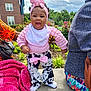 baby, toddler, child, outdoor, smiling, pink_headband, pink_top, cow_print_pants, bow, concrete, hand_support, plants, flowers, blanket, adult_hand, cloudy_sky, building, grass, person, colorful_clothing