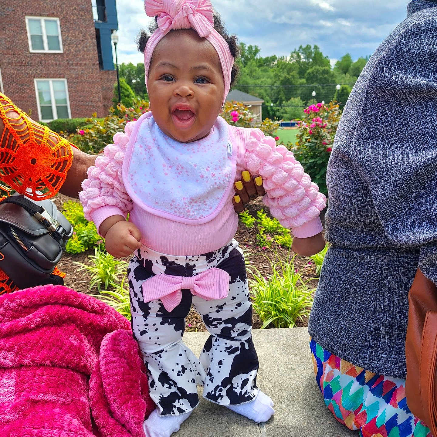 Shawty-Zuri joined the competition — help win amazing prizes! adult_hand, baby, blanket, bow, building, child, cloudy_sky, colorful_clothing, concrete, cow_print_pants, flowers, grass, hand_support, outdoor, person, pink_headband, pink_top, plants, smiling, toddler